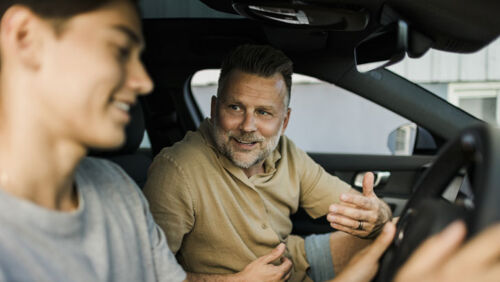 Father and adult son discussing a used car at a dealership before negotiating a purchase.